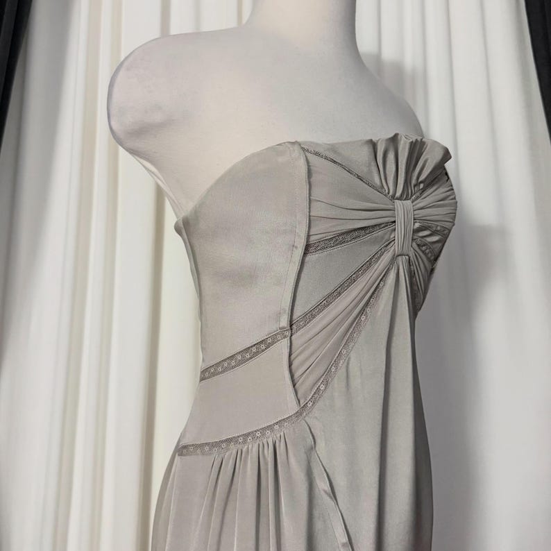 May include: A strapless light gray dress featuring a gathered bodice with a central vertical pleat detail. The garment includes diagonal panels accented with delicate lace trim along the seams, creating a structured yet fluid silhouette.