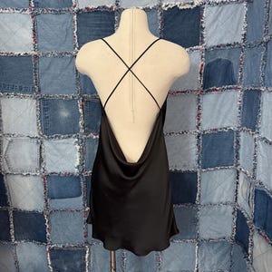 May include: A black satin slip dress with a cowl neckline and crisscross straps on the back. The dress is displayed on a mannequin against a patchwork denim background. The dress has a short hemline.