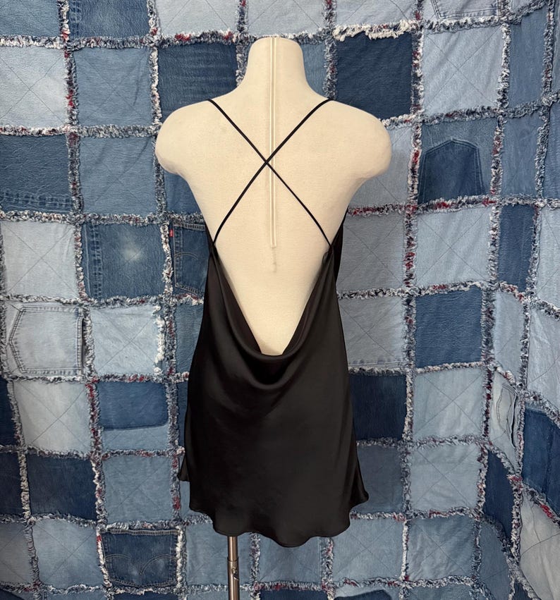 May include: A black satin slip dress with a cowl neckline and crisscross straps on the back. The dress is displayed on a mannequin against a patchwork denim background. The dress has a short hemline.