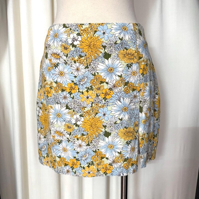 May include: A floral print mini skirt featuring a vibrant pattern of yellow, white, and light blue flowers on a white background. The skirt has a high waist and a slit on the side, showcasing a summery style.