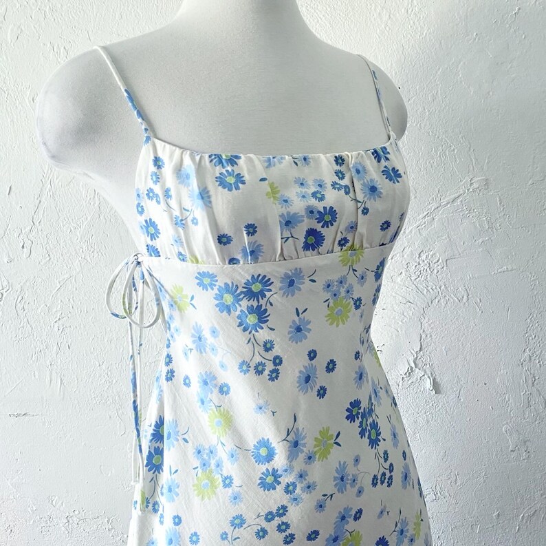 May include: A white sundress with a blue and green floral print. The dress has a square neckline and thin straps. The dress has a tie at the side.
