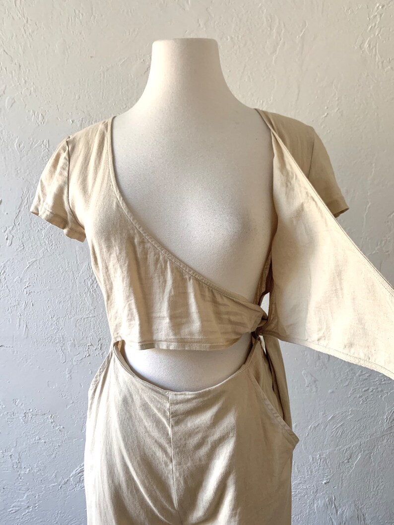 May include: A beige jumpsuit with a short-sleeved, wrap-style top and a cutout detail at the waist. The top has a tie detail at the side.