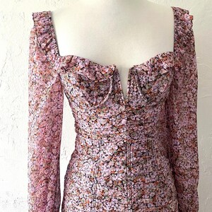 Op de afbeelding: Een roze jurk met bloemenprint, lange mouwen en een diep decolleté. De jurk heeft een aansluitend bovenlijf en een zwierige rok.