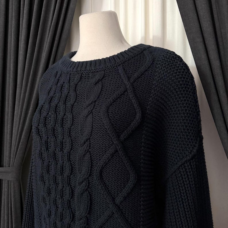 May include: A navy blue cable-knit sweater displayed on a mannequin. The sweater features a crew neck and intricate cable patterns across the chest and sleeves. The knitwear has a textured appearance and a classic, timeless design.