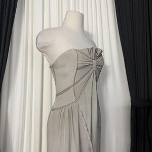 May include: A strapless light gray dress featuring a gathered bodice with a central vertical pleat detail. The garment includes diagonal seam lines accented with delicate lace trim, creating a structured silhouette that flows into a smooth skirt.