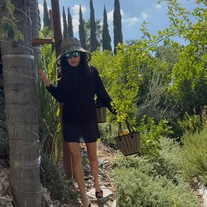 Puede incluir: Una persona con un vestido negro y un sombrero de sol de cuadros vichy lleva dos peque&ntilde;os bolsos de mano de cuadros vichy. Lleva gafas de sol negras y sandalias de tac&oacute;n negras. El fondo presenta exuberante vegetaci&oacute;n, &aacute;rboles y un cielo azul.