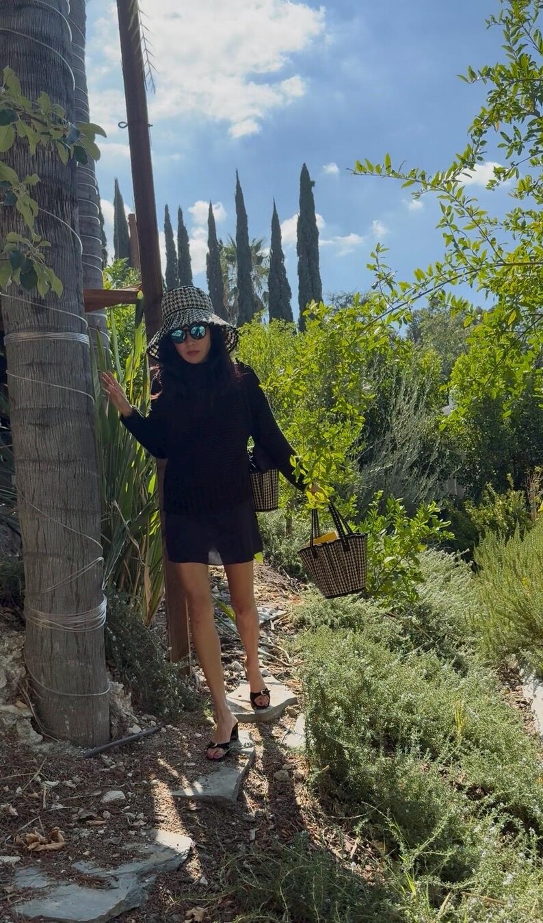 Puede incluir: Una persona con un vestido negro y un sombrero de sol de cuadros vichy lleva dos peque&ntilde;os bolsos de mano de cuadros vichy. Lleva gafas de sol negras y sandalias de tac&oacute;n negras. El fondo presenta exuberante vegetaci&oacute;n, &aacute;rboles y un cielo azul.