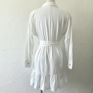 Puede incluir: Vestido blanco de manga larga con un dobladillo con volantes y un cintur&oacute;n de lazo en la cintura.