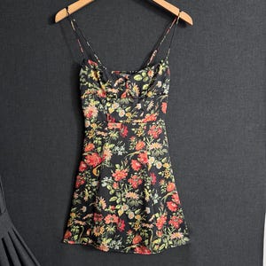 May include: A black floral print dress with spaghetti straps hangs on a wooden hanger. The dress features a vibrant pattern of red, yellow, and green flowers. The dress is a mini length.