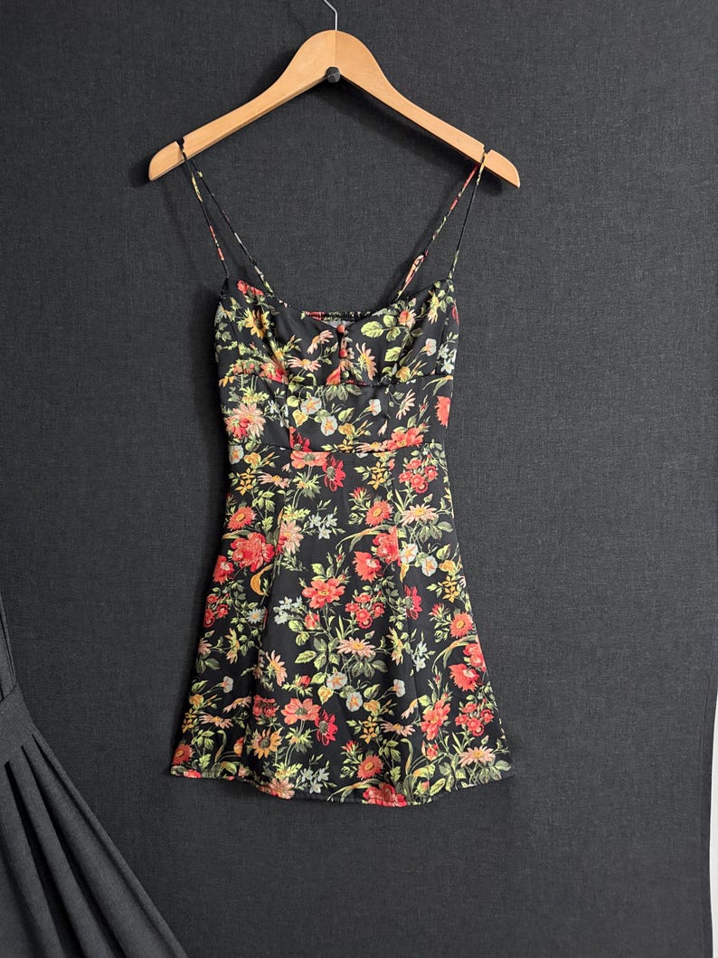 May include: A black floral print dress with spaghetti straps hangs on a wooden hanger. The dress features a vibrant pattern of red, yellow, and green flowers. The dress is a mini length.