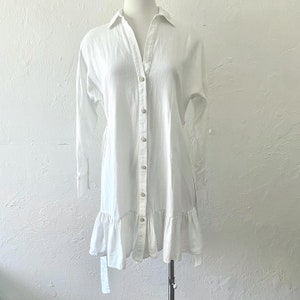 Puede incluir: Un vestido blanco de camisa de lino con un dobladillo con volantes y cierres de corbata en las mangas. El vestido tiene un ajuste holgado y un cuello en V.