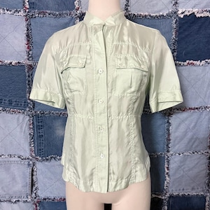 May include: Light green short-sleeved shirt with a mandarin collar and two chest pockets. The shirt has a fitted waist and button-up front. The fabric has a subtle sheen. The shirt is displayed on a mannequin.