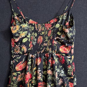 May include: A black floral print dress with spaghetti straps. The dress features a vibrant pattern of red, orange, yellow, and green flowers and leaves. The bodice has a smocked panel in the center. The dress is made of a lightweight fabric.
