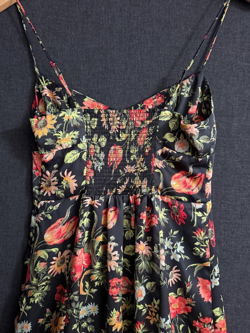 May include: A black floral print dress with spaghetti straps. The dress features a vibrant pattern of red, orange, yellow, and green flowers and leaves. The bodice has a smocked panel in the center. The dress is made of a lightweight fabric.