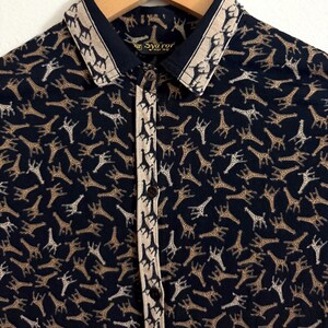 Op de afbeelding: Een zwart overhemd met knopen en een beige en bruine giraffenprint. Het overhemd heeft een beige kraag met een bruine bies en een gouden label met de tekst "Syaror" en "Made in Italy".