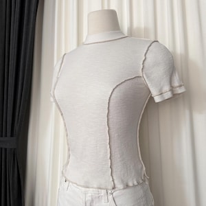 Puede incluir: Una camiseta blanca de manga corta con cuello alto. La camisa presenta costuras marrones visibles a lo largo de las costuras y los bordes, creando un elemento de diseño contrastante. La camiseta se muestra en un maniquí.