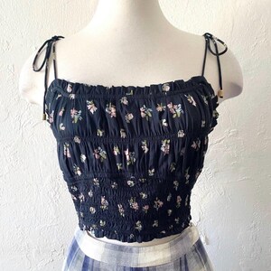 May include: A black crop top with a floral pattern. The top features a smocked bodice and tie straps. The floral design includes pink, green, and white flowers. The top is displayed on a mannequin.