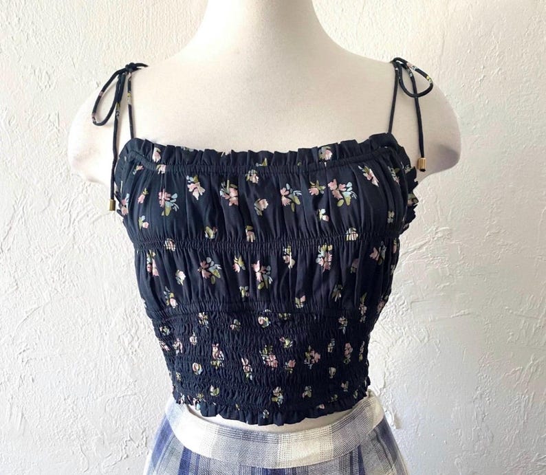 May include: A black crop top with a floral pattern. The top features a smocked bodice and tie straps. The floral design includes pink, green, and white flowers. The top is displayed on a mannequin.