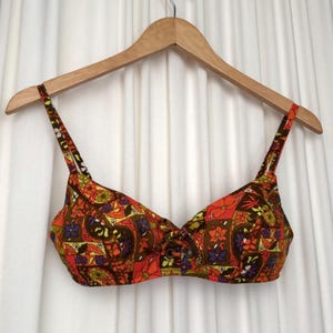May include: A vintage bikini top with a colourful floral pattern in orange, brown, yellow, and purple. The top is hanging from a wooden hanger against a white backdrop. The design features a square neckline and adjustable straps.