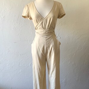 May include: A beige linen jumpsuit with a wrap-style bodice and short sleeves. The jumpsuit has a wide leg and a tie belt at the waist.