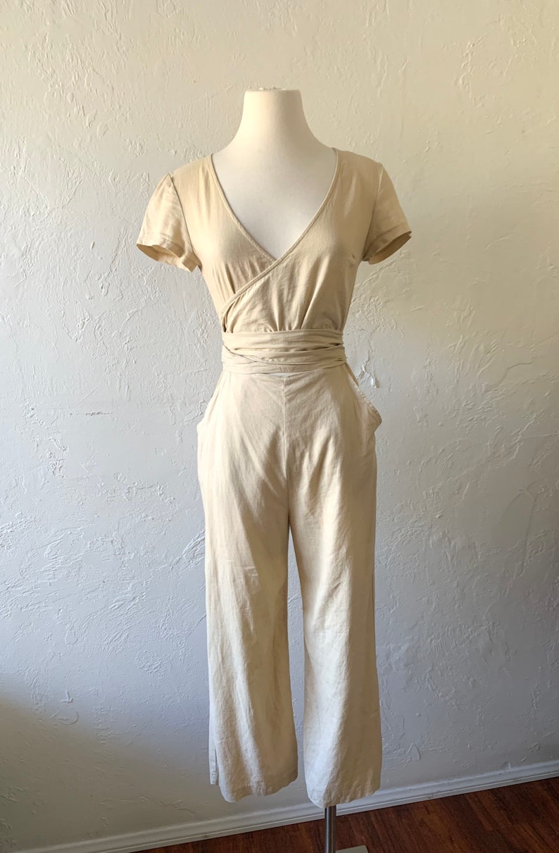 May include: A beige linen jumpsuit with a wrap-style bodice and short sleeves. The jumpsuit has a wide leg and a tie belt at the waist.
