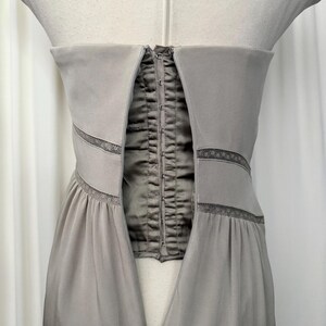 May include: A strapless taupe dress featuring a central corset-style panel with a vertical row of metal hook-and-eye closures. The bodice includes horizontal bands of delicate lace trim that extend across the front, transitioning into a gathered skirt.
