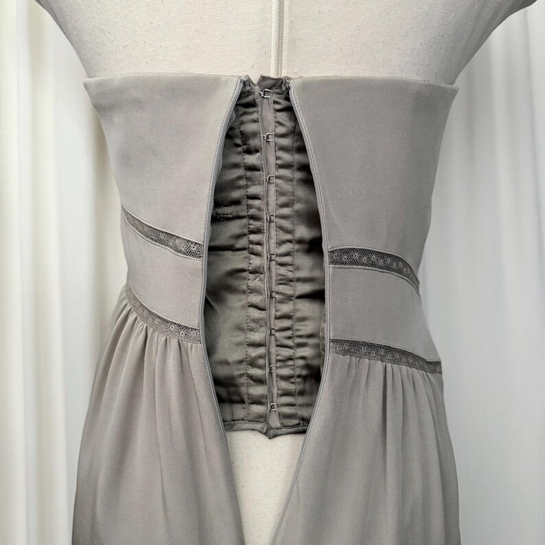 May include: A strapless taupe dress featuring a central corset-style panel with a vertical row of metal hook-and-eye closures. The bodice includes horizontal bands of delicate lace trim that extend across the front, transitioning into a gathered skirt.