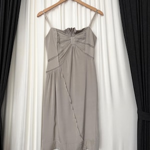 May include: A taupe-colored sleeveless dress featuring thin spaghetti straps, a gathered bodice with a central vertical detail, and delicate lace trim inserts. The skirt has a diagonal seam running from the waist to the hem, finished with a wide band.