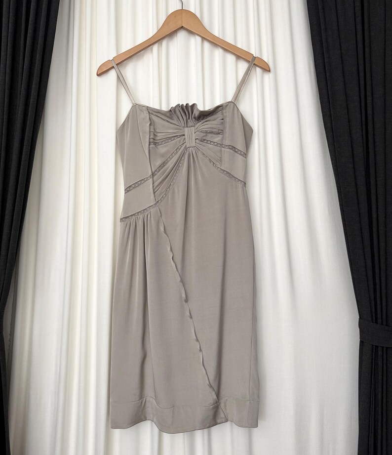 May include: A taupe-colored sleeveless dress featuring thin spaghetti straps, a gathered bodice with a central vertical detail, and delicate lace trim inserts. The skirt has a diagonal seam running from the waist to the hem, finished with a wide band.