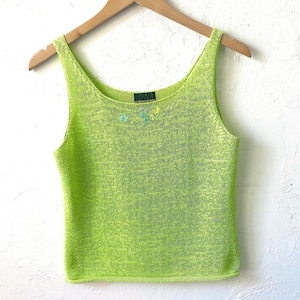 K&ouml;nnte beinhalten: Ein neongr&uuml;nes Strick-Tanktop mit Rundhalsausschnitt und kleiner floraler Verzierung am Halsausschnitt. Das &auml;rmellose Oberteil h&auml;ngt an einem Holzb&uuml;gel vor wei&szlig;em Hintergrund. Das Oberteil hat eine leuchtende, einfarbige Farbe.