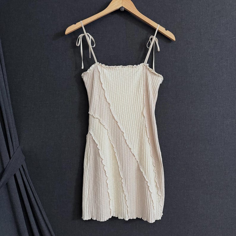 May include: A beige ribbed mini dress with tie straps, hanging on a wooden hanger against a dark gray background. The dress features a unique design with diagonal seams and a ruffled edge.