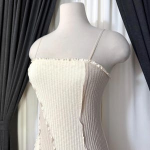 May include: A cream-colored ribbed knit top with thin spaghetti straps. The top features a contrasting panel of light brown fabric on the side. The neckline has a ruffled edge, and the garment is displayed on a mannequin.