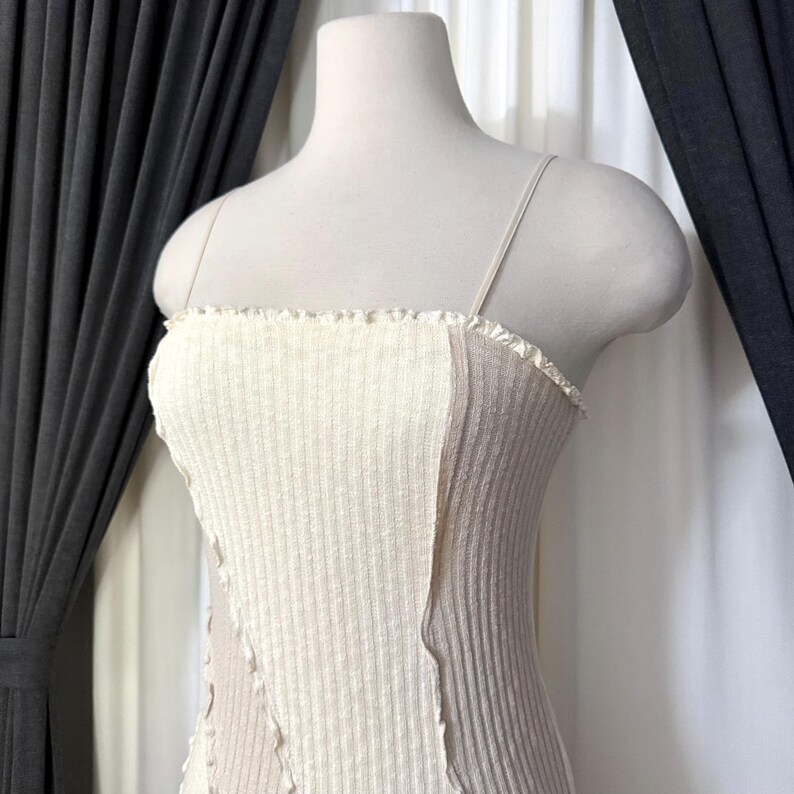 May include: A cream-colored ribbed knit top with thin spaghetti straps. The top features a contrasting panel of light brown fabric on the side. The neckline has a ruffled edge, and the garment is displayed on a mannequin.