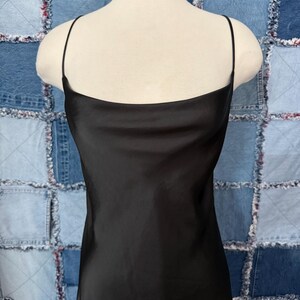 May include: A black slip dress with thin spaghetti straps and a square neckline. The dress is made of a shiny, silky material and is displayed on a mannequin. The background is a patchwork of denim squares.