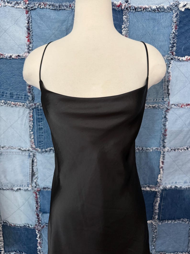 May include: A black slip dress with thin spaghetti straps and a square neckline. The dress is made of a shiny, silky material and is displayed on a mannequin. The background is a patchwork of denim squares.
