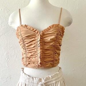 May include: A brown, ruched, cropped tank top with a sweetheart neckline and thin straps. The top has a ruffled trim around the neckline.