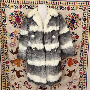 May include: A long, striped, gray and white fur coat with a large collar. The coat has a double breasted closure with buttons.