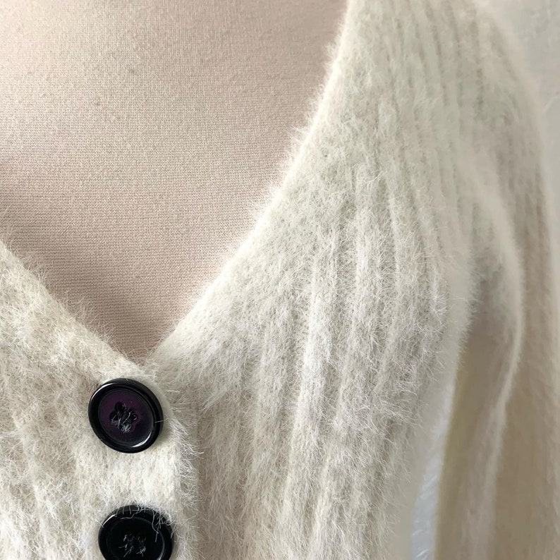 Peut inclure: Un cardigan blanc en fourrure avec un col en V et des boutons noirs.