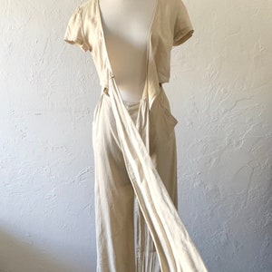 May include: A beige linen jumpsuit with a wrap-style top and a low back. The top has short sleeves and a V-neckline. The jumpsuit is tied at the waist with a long sash.