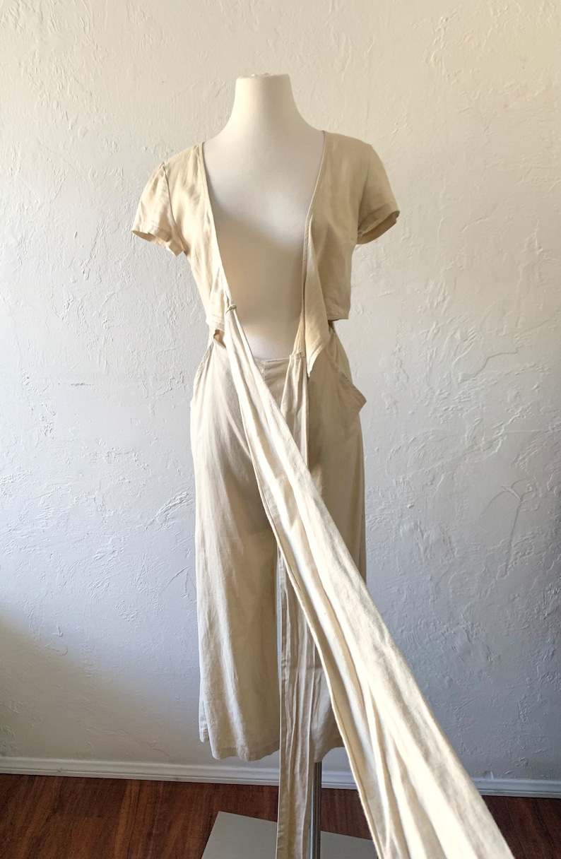 May include: A beige linen jumpsuit with a wrap-style top and a low back. The top has short sleeves and a V-neckline. The jumpsuit is tied at the waist with a long sash.