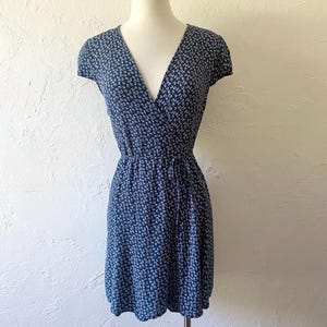 May include: A navy blue wrap dress with a white floral pattern. The dress features short sleeves, a V-neckline, and a tie waist. The dress is displayed on a mannequin against a white background.