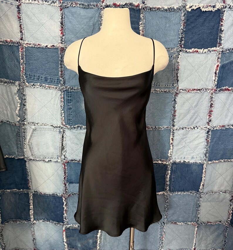 May include: A black slip dress with thin shoulder straps. The dress has a simple, elegant design and a slightly flared hemline. The dress is displayed on a mannequin against a patchwork denim background.