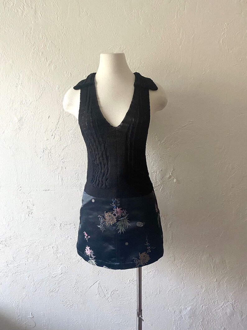 K&ouml;nnte beinhalten: Ein schwarzes &auml;rmelloses Strickoberteil mit V-Ausschnitt und einem schwarzen Rock mit Blumenmuster. Der Rock hat eine satinartige Textur.