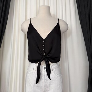May include: A black satin camisole top with a V-neckline and spaghetti straps. The top features a tie detail at the waist and three small buttons down the center. The top is displayed on a mannequin.
