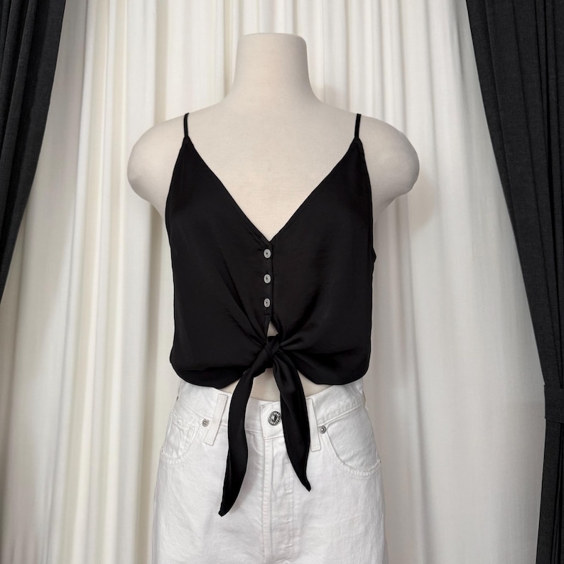 May include: A black satin camisole top with a V-neckline and spaghetti straps. The top features a tie detail at the waist and three small buttons down the center. The top is displayed on a mannequin.