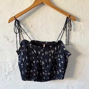May include: A navy blue crop top with a floral pattern, featuring adjustable spaghetti straps. The top is gathered and has a smocked bodice. It is hanging on a wooden hanger against a textured white wall.