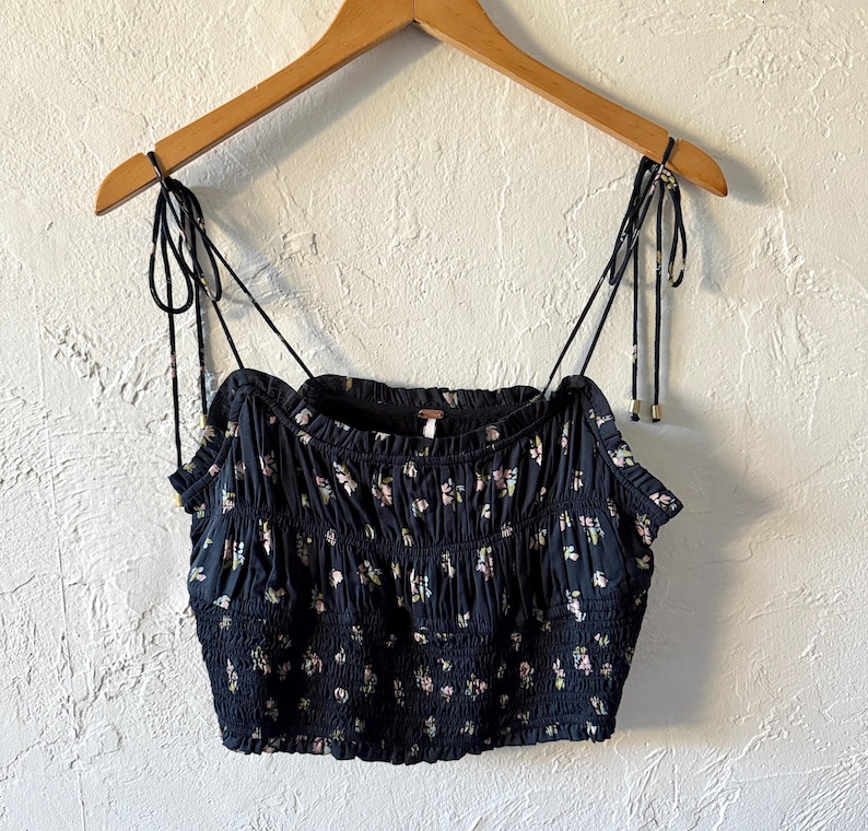 May include: A navy blue crop top with a floral pattern, featuring adjustable spaghetti straps. The top is gathered and has a smocked bodice. It is hanging on a wooden hanger against a textured white wall.
