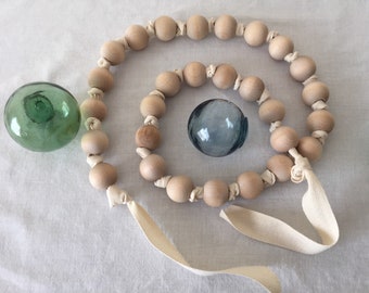 May include: A decorative wooden bead garland with a cream-colored ribbon, and two glass floats. The garland is made of light-colored wooden beads, connected by cream-colored fabric. One glass float is green, and the other is blue.