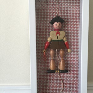 May include: A framed wooden marionette with a painted face, wearing a black hat, red sleeves, and a brown skirt. The marionette is suspended in a white frame against a red and white patterned background. A red ball hangs below.