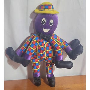 May include: A plush octopus toy with a purple head, wearing a yellow hat with a red and blue plaid pattern. The body features a vibrant red, blue, and yellow plaid design, a black bow tie, and eight tentacles ending in solid black fabric feet.